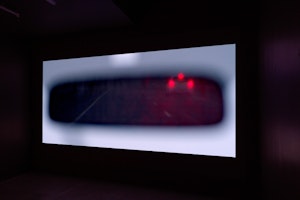 A film is projected onto a wall, in it we see the brake lights of a small car in a blurry rear-view mirror.