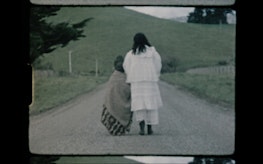 Two women in historic and traditional dress walk down a rural road. Visible film still.