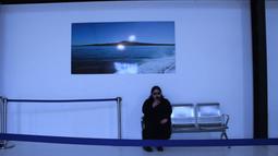 In a dim grey-blue lit room a person sits on an airplane lounge bench looking at their phone. A large photograph of an island is on the wall above them.