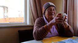 A person wearing a wool beanie and caridgan sits at a table by a window drinking a cup of tea.