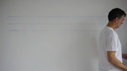 A person walks back and forth drawing thin blue lines on a white wall.