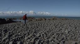 A man in a red and black flannel and beanie is walking along rocks at the coast. There are mountains and clouds in the distance.