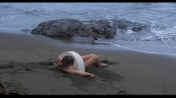 A person lies on sand by the waters edge, there is a semicircular sculpture over their torso.