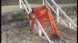 A person in an orange jumpsuit picks cleans a sandy beach, they are holding a rake.
