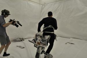 A room lined from floor to ceiling with white paper. At left, a camera operator is shooting a video of the artist Mark Harvey running towards a wall. Mark Harvey is wearing the tattered remains of a costume made entirely of newspaper. The video operator points their camera towards Harvey's feet, which are still covered in newspaper.