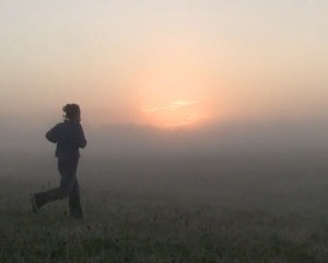 A person is jogging across a grassy field in the early morning. The landscape is covered in thick fog, blurring the horizon line. We can see the red glow from sunrise. The person running appears slightly mid-air, in between steps. The dawn makes them look like a silhouette on the land. The grass is covered in dew or frost.