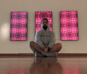 A person in their 20's sits in front of three neon lit pink sculptures which are hung on the wall. The panels look like digital versions of the traditional Māori tukutuku panel