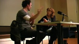 Two people sit at a desk on a stage having a conversation. There is a microphone in front of them.