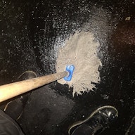 A hairy mop is being pushed against a shiny black concrete floor in a sex on site club. The image is taken looking down at the mop and the artists feet, mid way through mopping the floor.