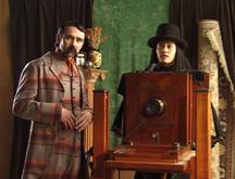 In a colonial photographers studio a comically mannered British gentleman and a Māori photographer stand by a large format camera