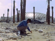 A person bends down amongst sand and rocks, there are palm trees and a large dome-topped building behind them.