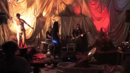 A band performs in a red and gold lit room, a naked man throws something from the corner.