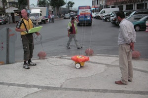 A street scene with people gathered round a small yellow object with wheels that is attached to four thin strings.