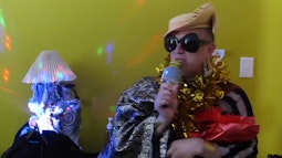 A person is singing into a gold microphone while wearing gold tinsel. There is a colourful party light shining beside them.