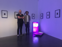 Steve and Christian stand in a gallery which displays small photographs in large frames neatly on the walls, two tvs rest against the wall and the floor of the gallery displaying a bright, pink image