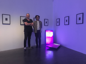 Steve and Christian stand in a gallery which displays small photographs in large frames neatly on the walls, two tvs rest against the wall and the floor of the gallery displaying a bright, pink image