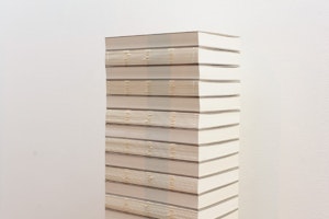 A tall stack of books that have their spines removed sits neatly in front of a white wall