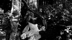 A person sits on the ground surrounded by bush while holding a white sheet.