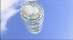 Two white ping pong balls sit inside a glass jar upon a mirror outside, the sky is reflected in the mirror and jar.