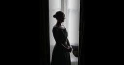A person stares contemplatively out a window between large dark curtains.