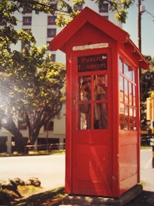A scanned image of a old photo of a person standing in a red phone box, holding a phone to their head.