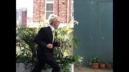 A person wearing a black suit runs across the frame. There are potted plants behind them.