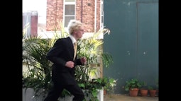 A person wearing a black suit runs across the frame. There are potted plants behind them.