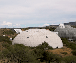 A giant white biodome with triangular tiles set in a grassy landscape under a  blue and cloudy sky.
