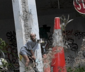 The artist leans against a tall concrete poll whilst a phone on a tripod is filming them. They are in an abandoned car park with wet concrete and weeds.
