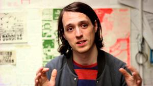 A person with straight, shoulder length brown hair is gestating with their hands. They are wearing a red top and casual grey jacket. Behind them is some blurred art work as if this photo has been taken in a studio or gallery. They look beyond the camera as if they are speaking to somebody standing next to it. They appear to be in mid-converstaion explaining something. 