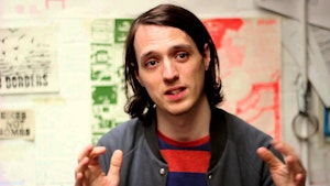 A person with straight, shoulder length brown hair is gestating with their hands. They are wearing a red top and casual grey jacket. Behind them is some blurred art work as if this photo has been taken in a studio or gallery. They look beyond the camera as if they are speaking to somebody standing next to it. They appear to be in mid-converstaion explaining something. 