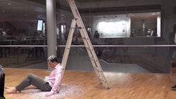 A man sits at the bottom of a ladder being covered in paper dust as a part of a performance.