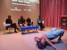 Four people sit a panel in the background whilst a performer in the foreground stretched their body across the floor