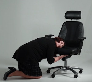 Claire wears a pencil skirt, blazer and high heels whilst tenderly hugging an office chair, resting her face against the seat