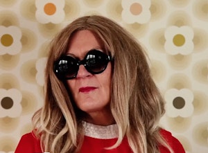 A person wearing large dark glasses, red lipstick, and a blonder wig. They stand in front of a 1970s-style wallpaper.