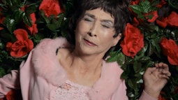 An elderly woman wearing a pink fluffy shawl lies in a bed of fake roses.