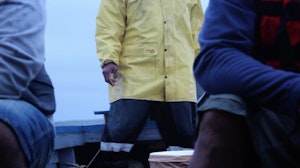 A sailor wearing oilskins stands in the stern of a small boat