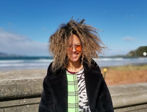 A photo of Israel Randell standing facing the camera. She wears sunglasses and her curly hair is blown by the wind. Behind her is the ocean.