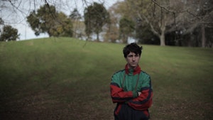 A young person stands in a park with their arms folded wearing a sports jacket