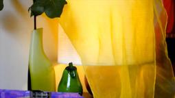 A yellow piece of fabric is draped over a lamp. There is a green vase with a rose beside the lamp.