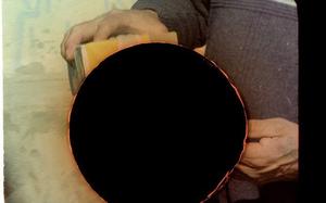 An aged photo of hands performing an action is obscured by a large hole in the image. The hole looks as if it occurred within analogue photographic processes as if it is acidic.