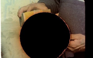 An aged photo of hands performing an action is obscured by a large hole in the image. The hole looks as if it occurred within analogue photographic processes as if it is acidic.