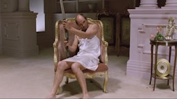 A man sits in a gold gilded chair in a white slip dress holding a shotgun.