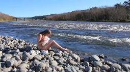 A person sits naked on river-side rocks while holding a crumpled white sheet.