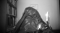 A person disguised in a rubbish bag holds a make up mirror to their face.