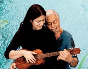 Kahurangiariki sits on their mother's lap, playing ukelele together. Behind them is a bird's eye view of water flowing