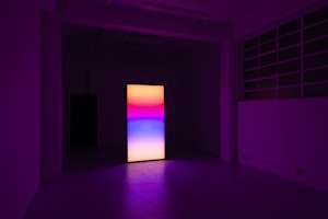 A bright screen illumanates a darkened gallery space. Gradient hues of orange, red, pink, blue bleed against one another slightly