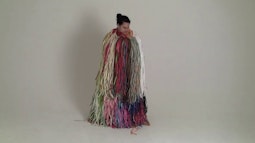 A person stands in a studio wearing a full length rainbow raffia outfit.
