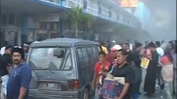 A large crowd of people riot on a street in Tonga, there is a building afire in the distance.