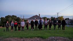 Fourteen people stand in a line on a lawn facing the camera. There is a house behind them.